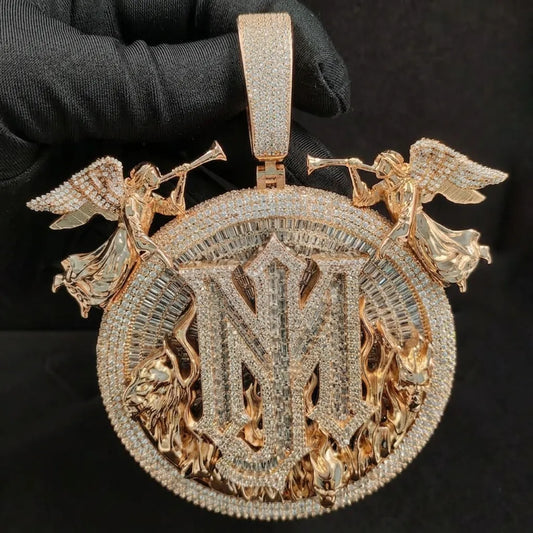A large round gold-toned pendant with a matching bail, heavily embellished with clear stones. The design features a central monogram-style letterform, angel figures on the sides holding trumpets, and flame motifs near the bottom, all shown against a black background with a gloved hand holding it.