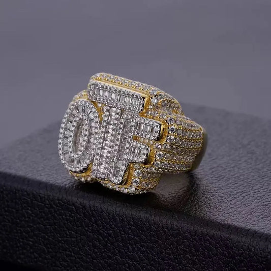 Two tone yellow gold and silver plated OTF bubble letter ring, full pave baguette and round moissanite stones, bold mens hip hop icy ring resting on textured black leather surface