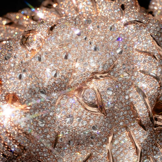 A close-up of a highly embellished rose gold jewelry piece covered in sparkling clear crystals and pavé stones, with a prominent central round gemstone and organic metallic rose gold settings.