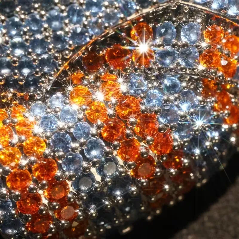Close-up of a rhinestone-covered curved surface with alternating rows of silver-blue and amber-orange faceted gems set in silver-toned metal, with bright sparkle highlights.