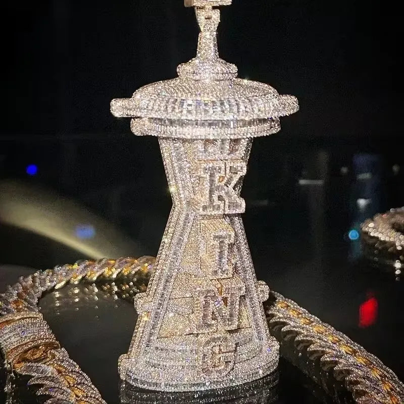 A heavily embellished silver-toned jewelry piece, likely a necklace or pendant, covered in sparkling clear stones and featuring the word 'KING' vertically. It is displayed on a dark reflective surface with gold-toned chains in the foreground.