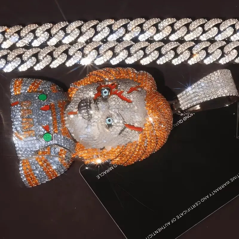 A heavily embellished jewelry piece featuring a silver-toned face with orange rhinestone hair, blue gemstone eyes, and green gem accents, displayed with a matching silver chain on a black surface.
