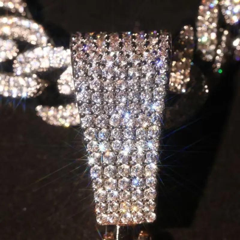 A close-up of a silver-toned jewelry piece, likely a brooch or decorative pin, densely covered with clear rhinestones and cubic zirconia stones, set against a dark background with blurred sparkling jewelry.