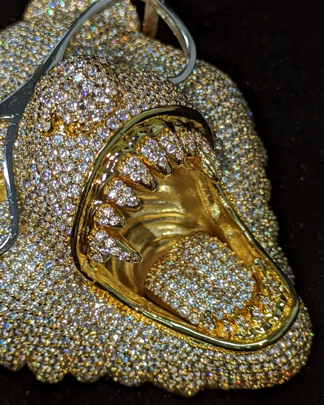 A close-up of a gold-toned shark mouth pendant or charm covered with sparkling clear stones, set against a dark background.