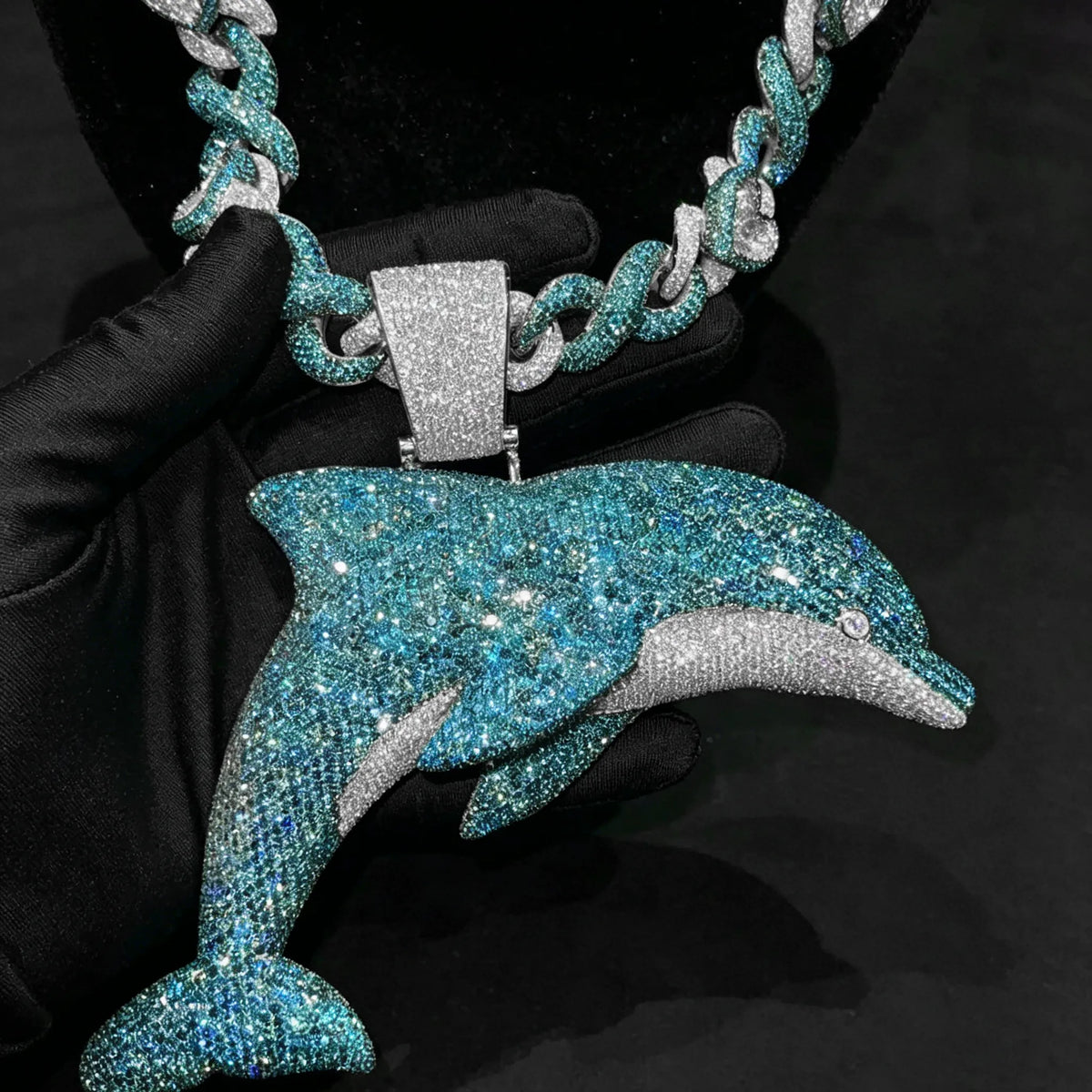 A glittery dolphin-shaped pendant necklace held by a black gloved hand against a dark background. The pendant and matching chain are covered in sparkling blue and silver rhinestones, with a silver-toned bail.
