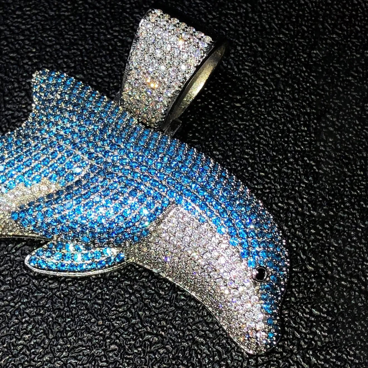 A dolphin-shaped pendant covered in blue and silver rhinestones, displayed on a textured black background.