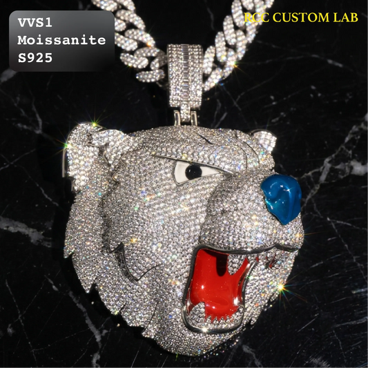 A large tiger head pendant necklace covered in sparkling silver stones, hanging from a thick silver chain. The pendant has a blue gemstone for a nose and a bright red gemstone for a mouth, set against a black marble background.