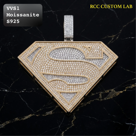 A Superman-style shield pendant with a gold-tone border and pavé-set stones, shown on a black marble background. Text in the top left reads 'VVS1 Moissanite S925' and text in the top right reads 'RCC CUSTOM LAB'.