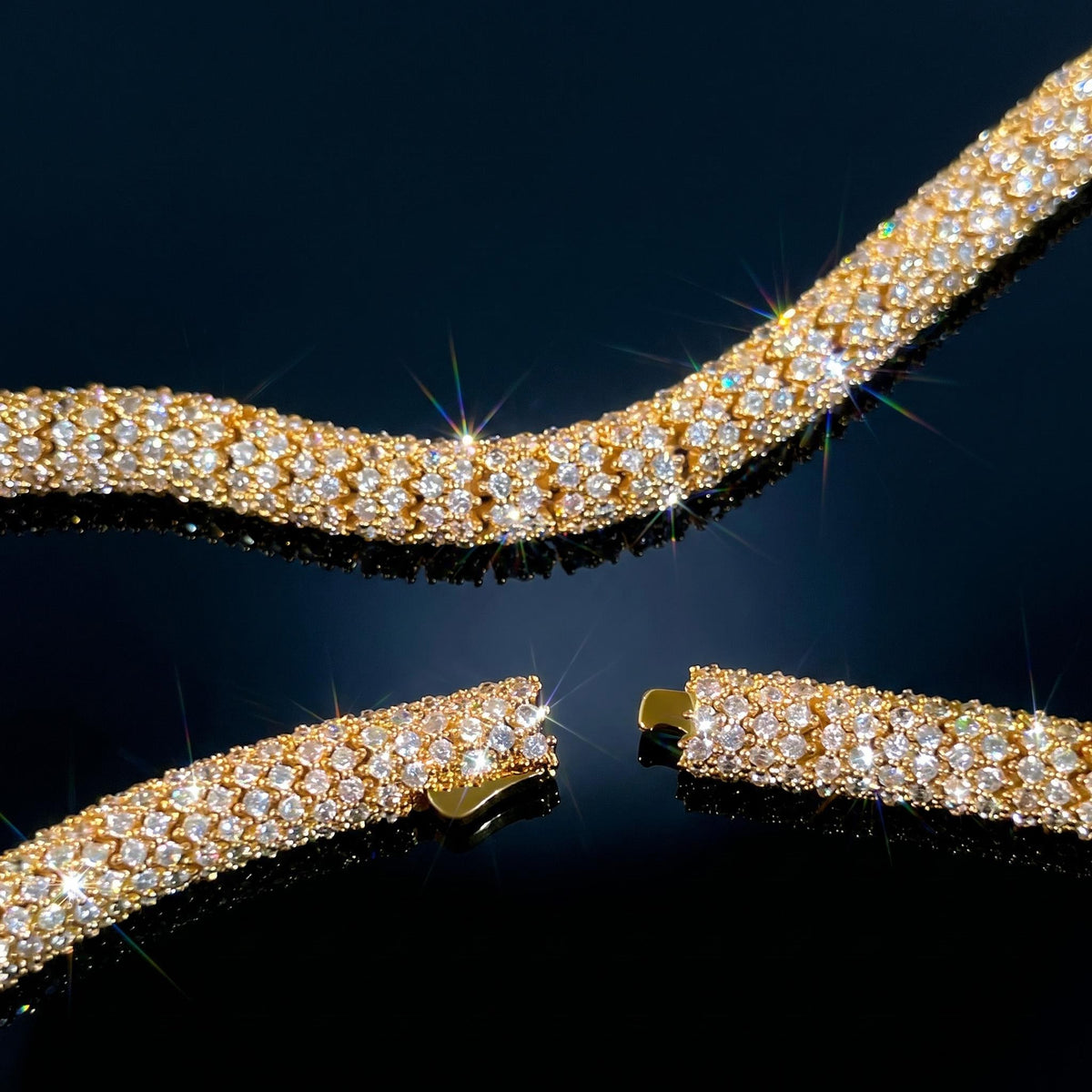 A gold-toned tennis bracelet with pavé-set clear stones, shown in multiple curved segments on a glossy black reflective surface. The clear stones are set in a yellow gold-tone metal.