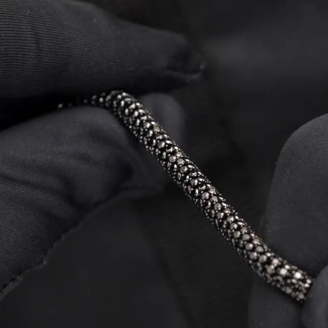 A close-up of a black chain bracelet with pavé-set clear stones, held between black-gloved hands against a dark background.