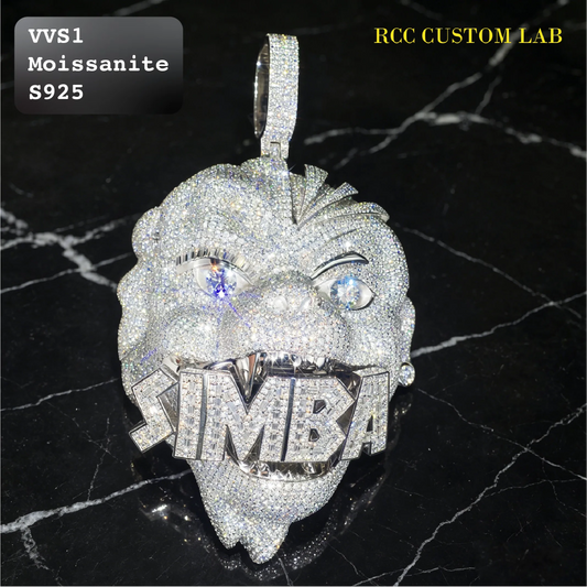 A lion head pendant covered in sparkling clear stones, featuring a blue gemstone for one eye and the word 'TIMBA' across its mouth. Shown on a black marble background with 'VVS1 Moissanite S925' and 'RCC CUSTOM LAB' text overlays.
