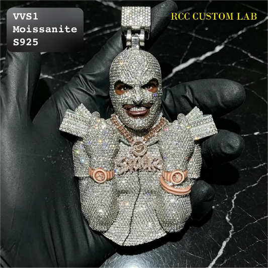 A heavily embellished custom pendant or charm shaped like a stylized face and upper body, covered in sparkling clear stones, held in a black gloved hand. The pendant includes rose-gold-toned circular details on the shoulders and chest.