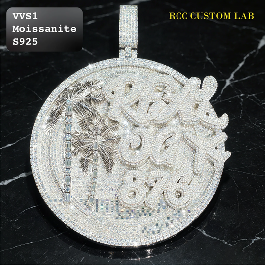 A large round custom pendant covered in sparkling clear stones, featuring an engraved design with palm trees and custom lettering. The pendant is displayed on a black marble surface, with text overlays reading 'VVS1 Moissanite S925' and 'RCC CUSTOM LAB'.