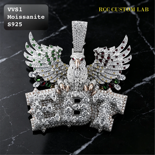 A large custom pendant featuring a silver eagle with spread wings and the letters 'EB' in front, heavily embellished with clear rhinestones. The pendant is displayed on a black marble background with text overlays reading 'VVS1 Moissanite S925' and 'RCC CUSTOM LAB'.