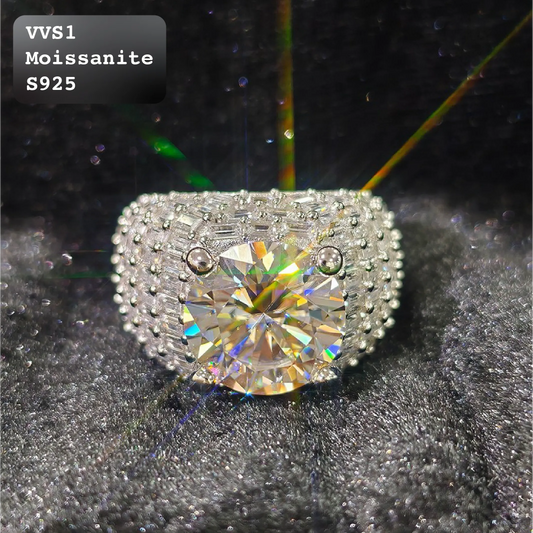 A silver-colored ring featuring a large round brilliant-cut moissanite center stone and a halo of smaller round and baguette-cut stones. The ring is displayed on a textured black background with a text overlay in the top-left corner that reads 'VVS1 Moissanite S925'.