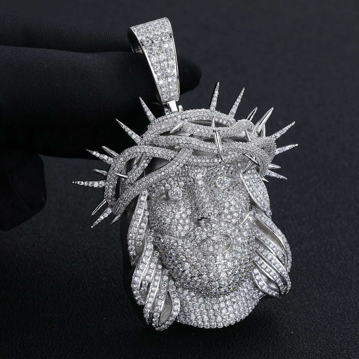 Close-up detail shot of fully paved moissanite Jesus face pendant in silver finish, held by black gloved hand, showcasing intricate crown of thorns detailing and micro-pave stone setting