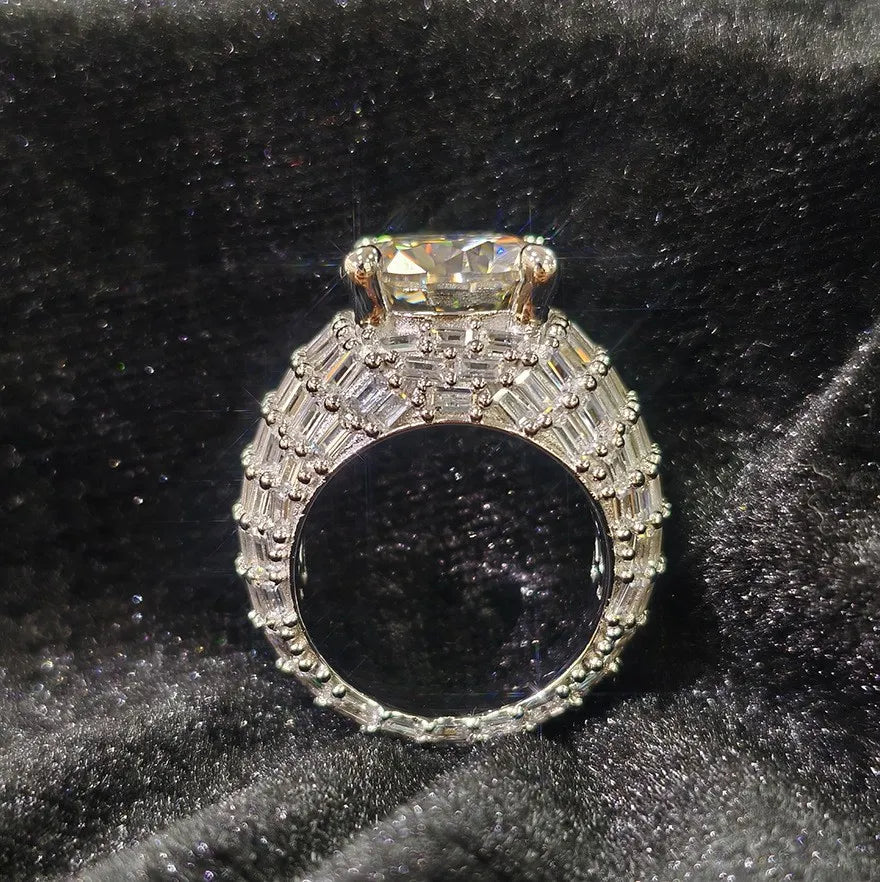 Side profile view of silver men's wide band moissanite ring, large round center stone secured by four prongs, band paved with baguette diamonds, hip hop bling jewelry on black velvet