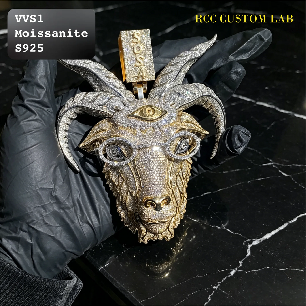 A custom moissanite and gold ram head pendant or charm held by a gloved hand on a black marble surface. The pendant features a detailed ram head face, curved horns, a single eye motif, and a hanging tag engraved with 'SOX'.