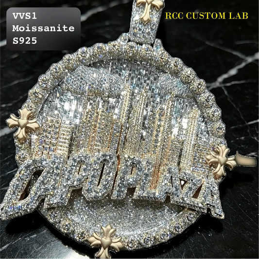 A close-up of a heavily embellished circular pendant or medallion with large clear stones, gold-toned lettering, and decorative floral accents on a dark marble-like surface. Text overlays read 'VVS1 Moissanite S925' and 'RCC CUSTOM LAB'.