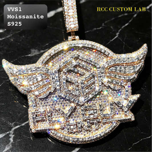A large winged pendant covered in sparkling clear rhinestones and cubic zirconia, displayed on a black marble background. The pendant features a central geometric design with the text 'VVS1 Moissanite S925' on a black overlay in the top left, and 'RCC CUSTOM LAB' in yellow text in the top right.