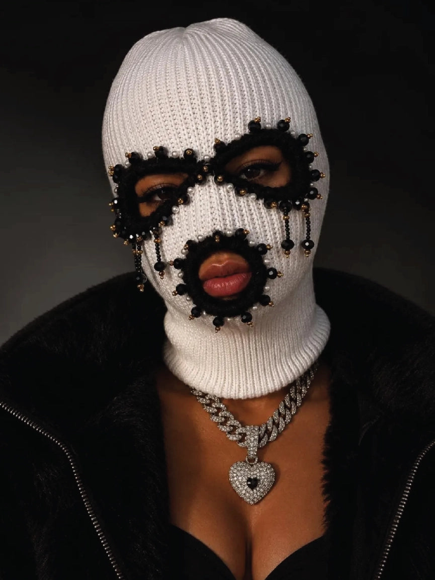 Female model portrait wearing white rib knit balaclava decorated with black beads, pearls and dangling tassels, paired with black fur coat and iced heart cuban chain, luxury streetwear fashion dark background